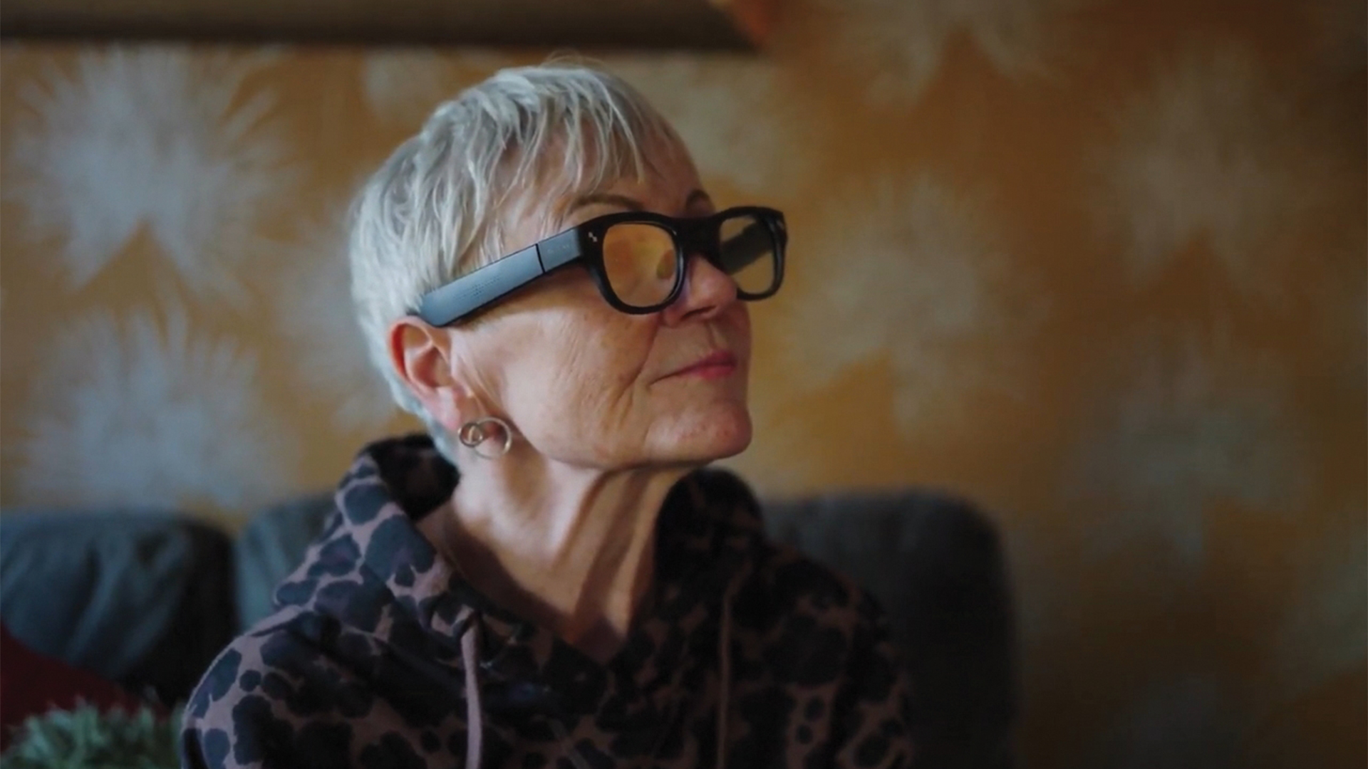 An older person wearing smart glasses sits in a warmly lit room.