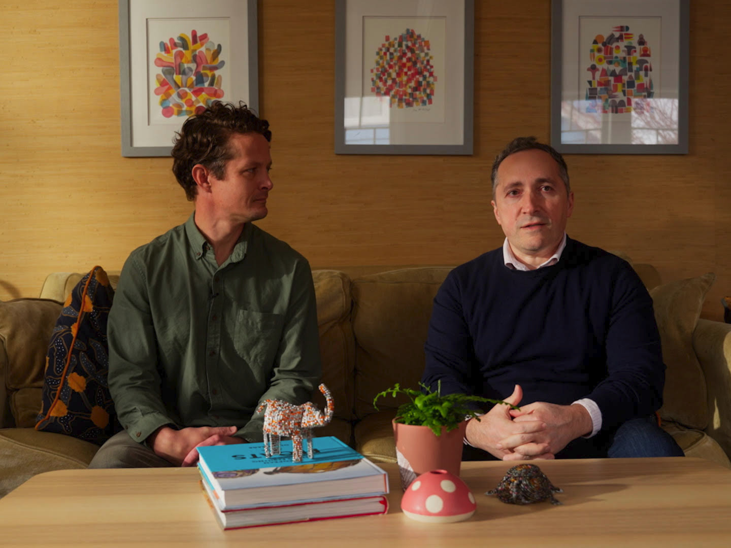Two NewDays founders sit together on a sofa behind a table with books and small objects.