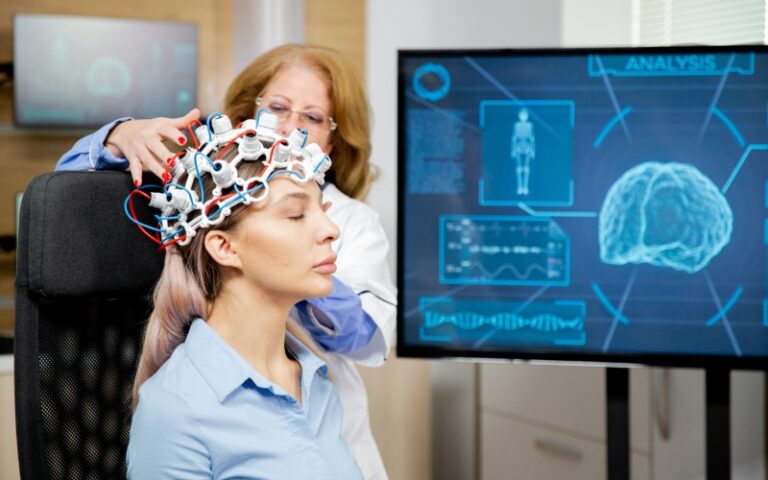 A clinician positions an EEG cap on a seated woman beside a monitor displaying brain-analysis graphics.